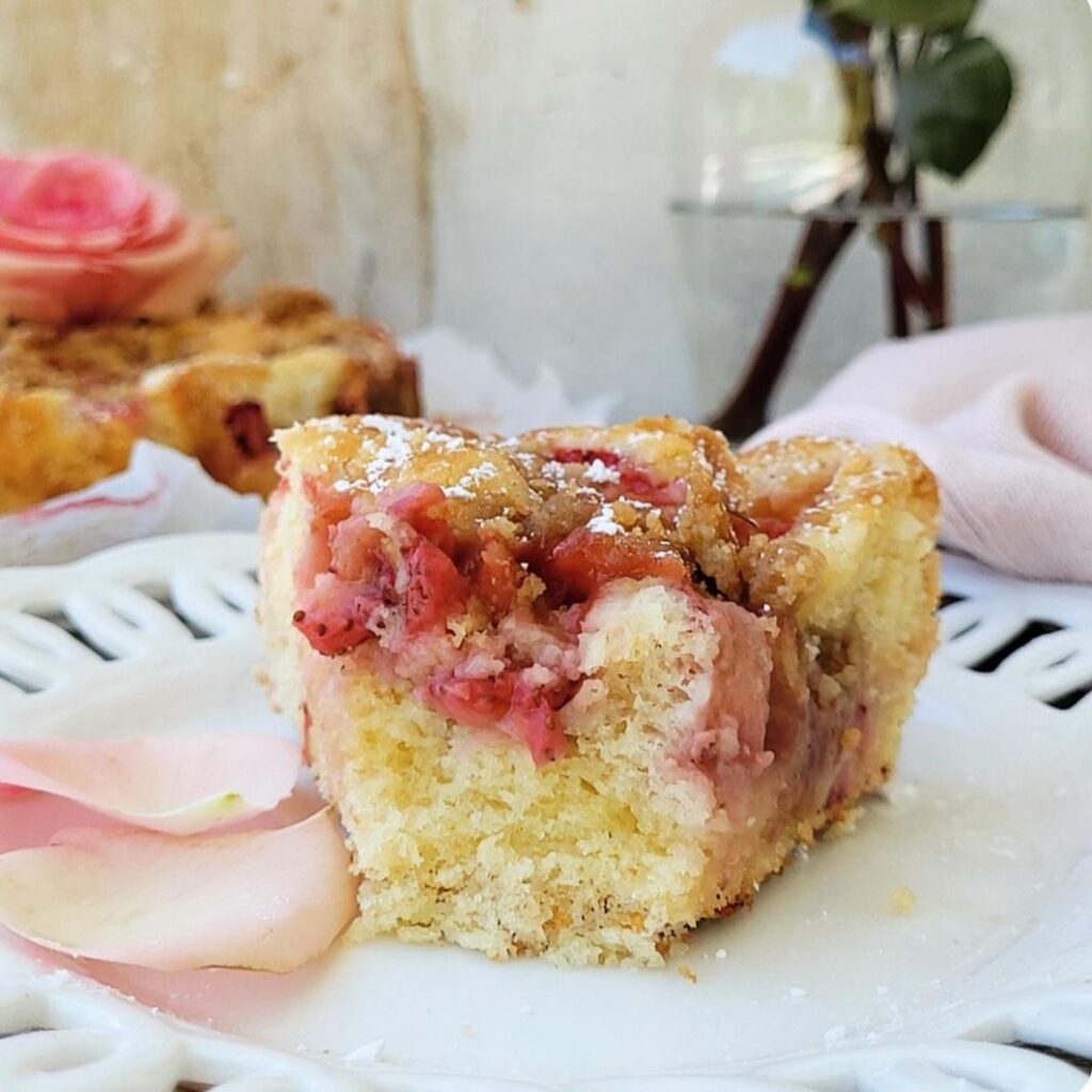 strawberry crumb cake. close up view of a slice on a white plate so you can see the inner crumb and strawberries. plate is styled with pale pink rose petals.  