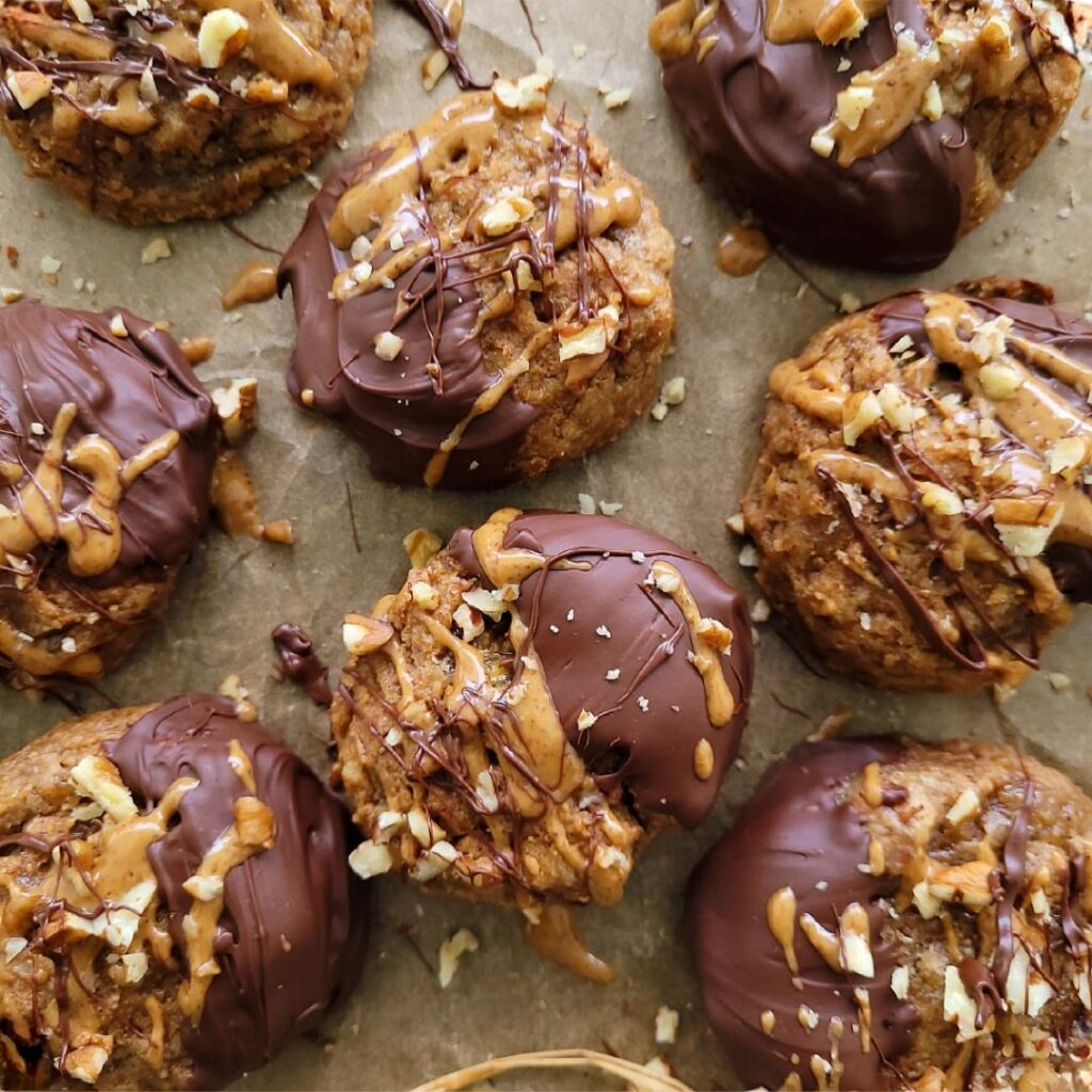 top down view of banana almond butter cookies on a baking sheet. cookies are topped with melted chocolate, drizzled with almond butter and garnished with pecans. 