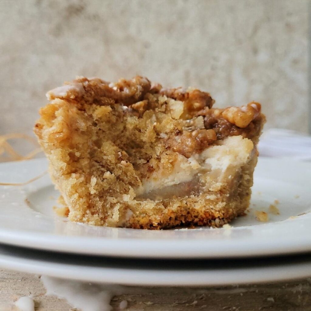 side view of a bitten slice of moist banana coffee cake on a white plate so you can see the walnut streusel topping, the inner crumb and the swirl of cream cheese in the middle. 