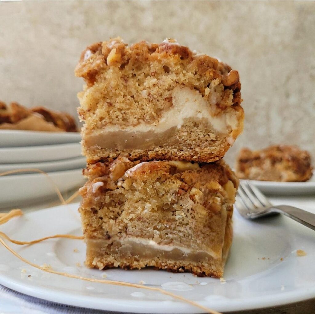 banana coffee cake swirled with cream cheese and topped with walnut streusel. side view image of two slices of coffee cake stacked on top of each other on a white plate so you can see the inner crumb.