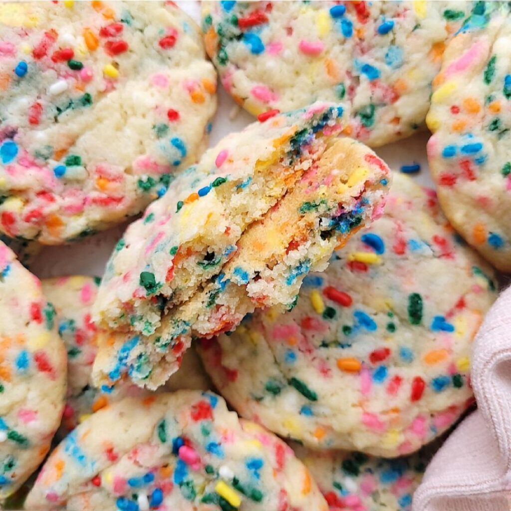 top down view of a pile of funfetti sprinkle sugar cookies. one cookie is broken in half and vertical so you can see the colorful inner crumb and rainbow sprinkles. 