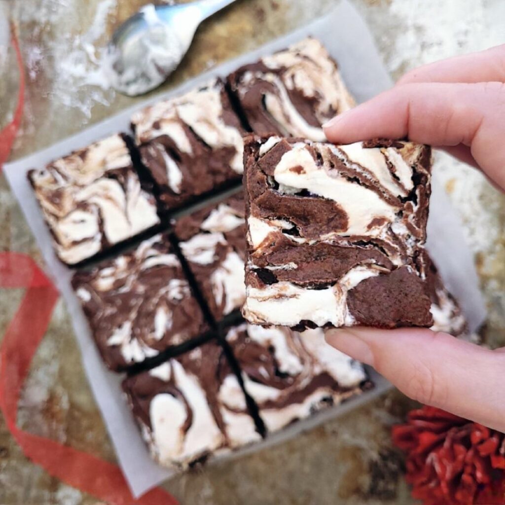 fudgy marshmallow brownies. close up view of a hand holding a brownie with marshmallow swirl. background is the remaining  pan of brownies cut into squares. 