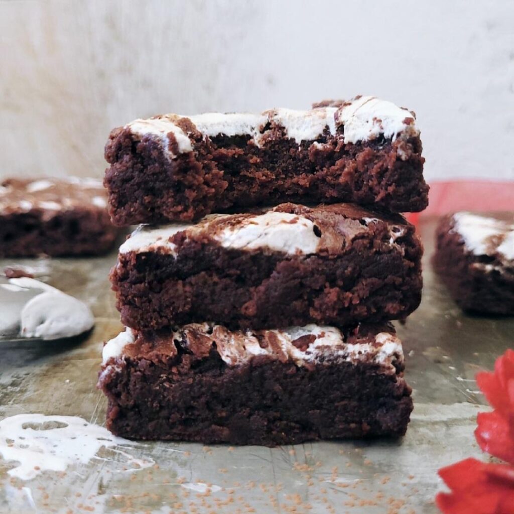 side view stack of three slices of chocolate marshmallow brownies. top slice has a bite missing so you can see the fudgy brownie inside and marshmallow topping. 