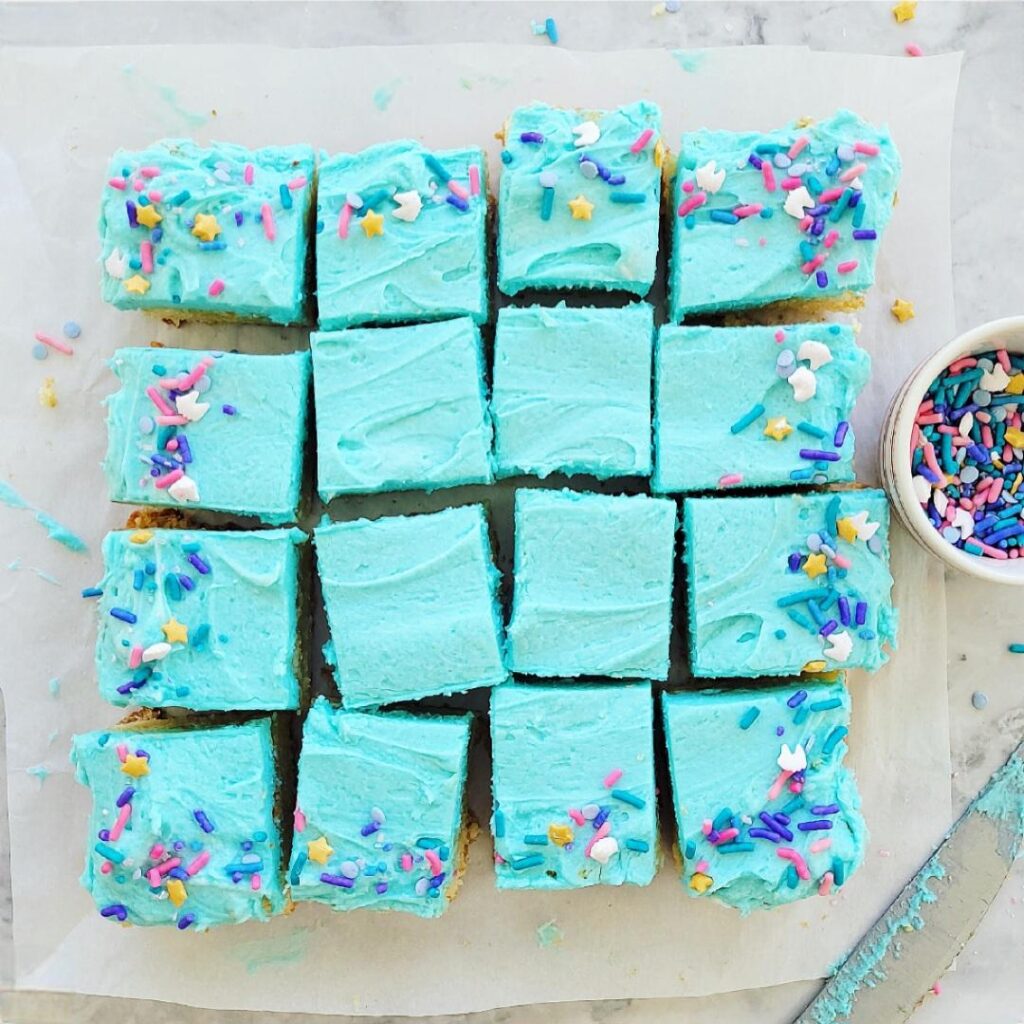 frosted sugar cookie bars cut into 16 squares. sugar cookie bars are frosted with blue buttercream and topped with colorful sprinkles. 