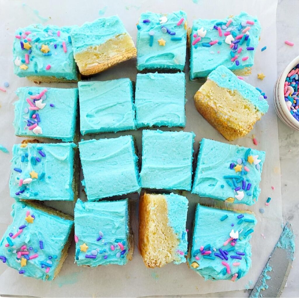 frosted sugar cookie bars. top down view of bars frosted with blue buttercream and topped with colorful sprinkles around the edges. three squares of the cookie bars are turned on their sides so you can see the inner crumb. 