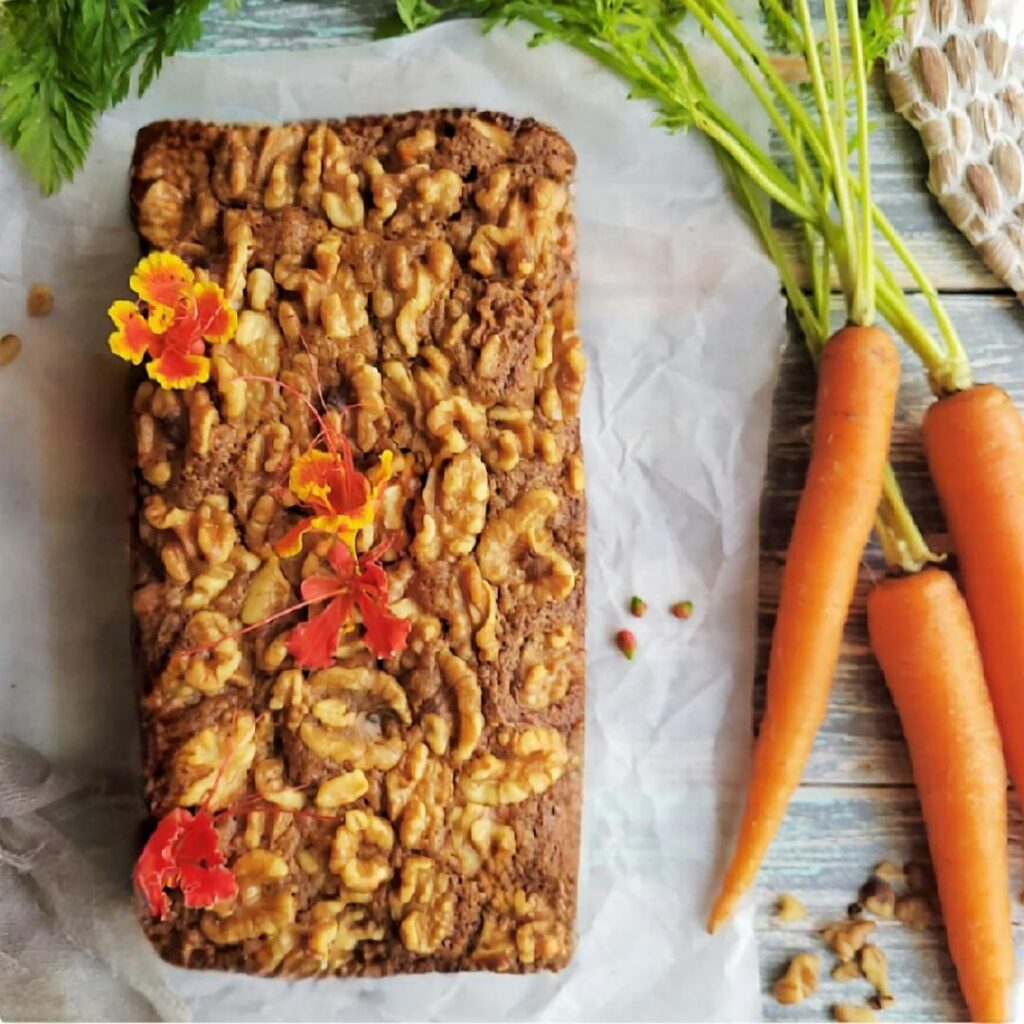 carrot bread topped with walnuts. Top down view of an uncut loaf of bread. image is styled with stemmed carrots, small red flowers and walnut pieces. 