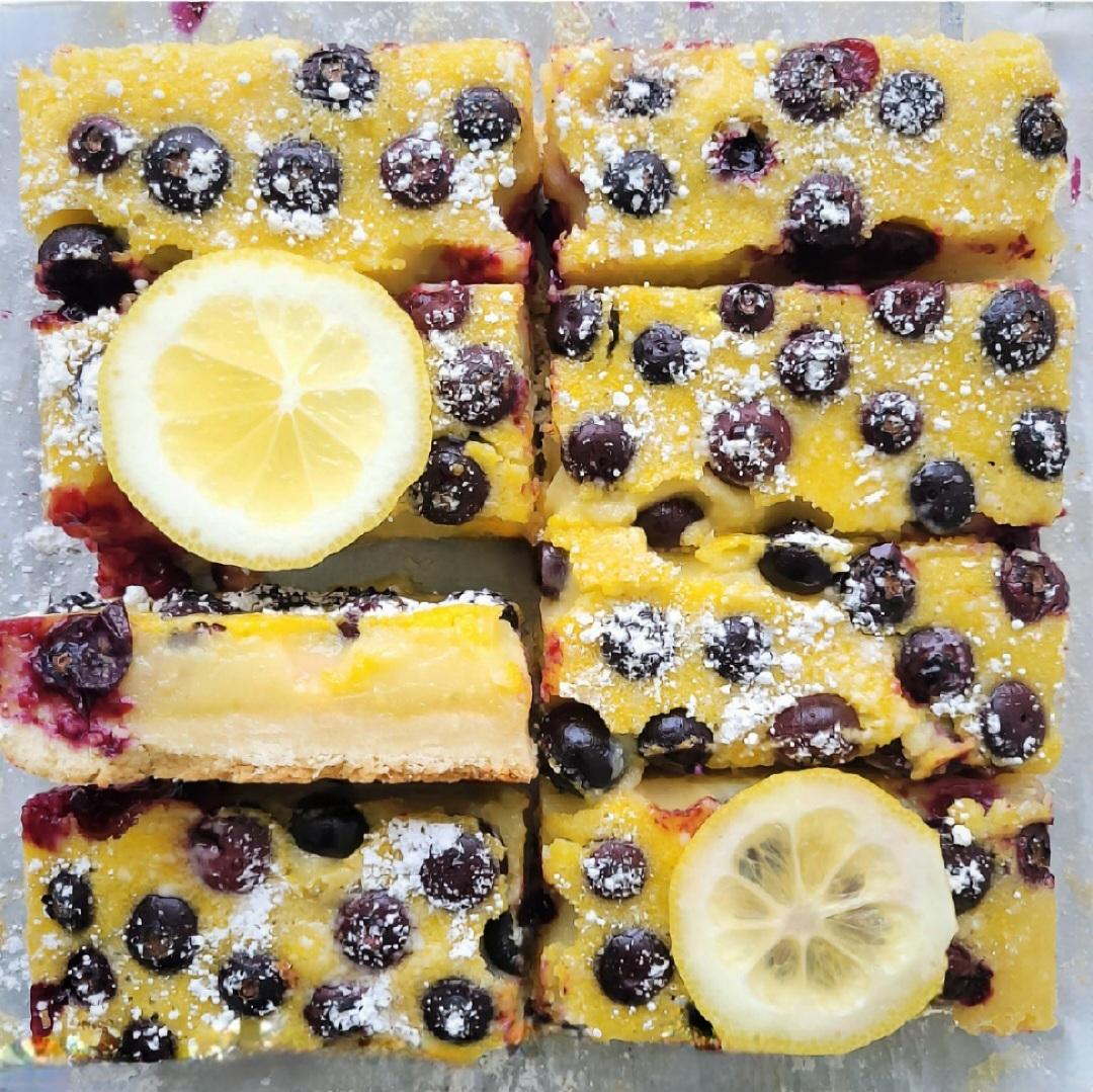 lemon shortbread bars with blueberries. top down view of sliced shortbread. one bar is turned on its side so you can see the layers of shortbread, lemon curd and blueberry topping. 