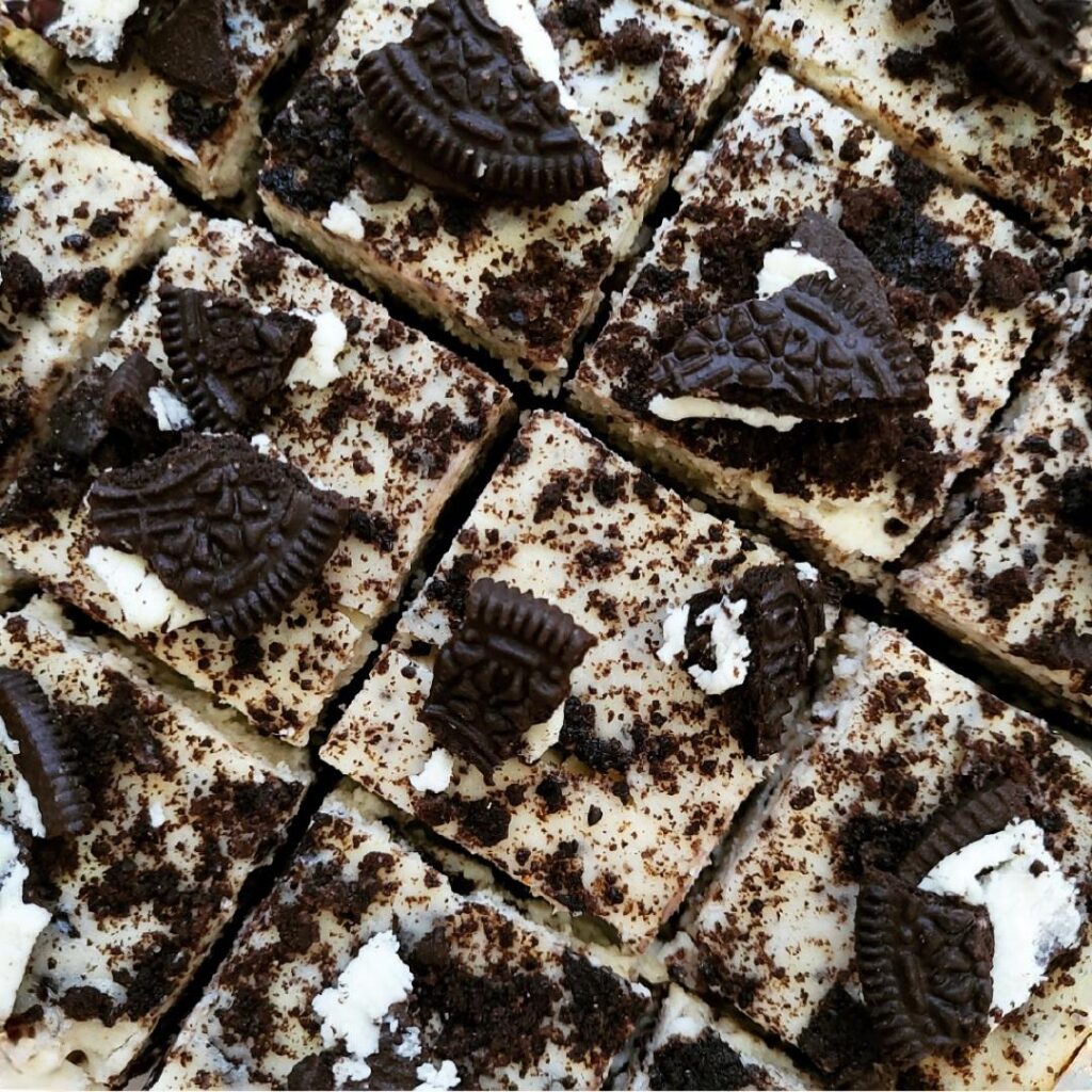 oreo cheesecake bars. top down view of a batch of cheesecake bars cut into squares and topped with broken Oreo cookeis. 