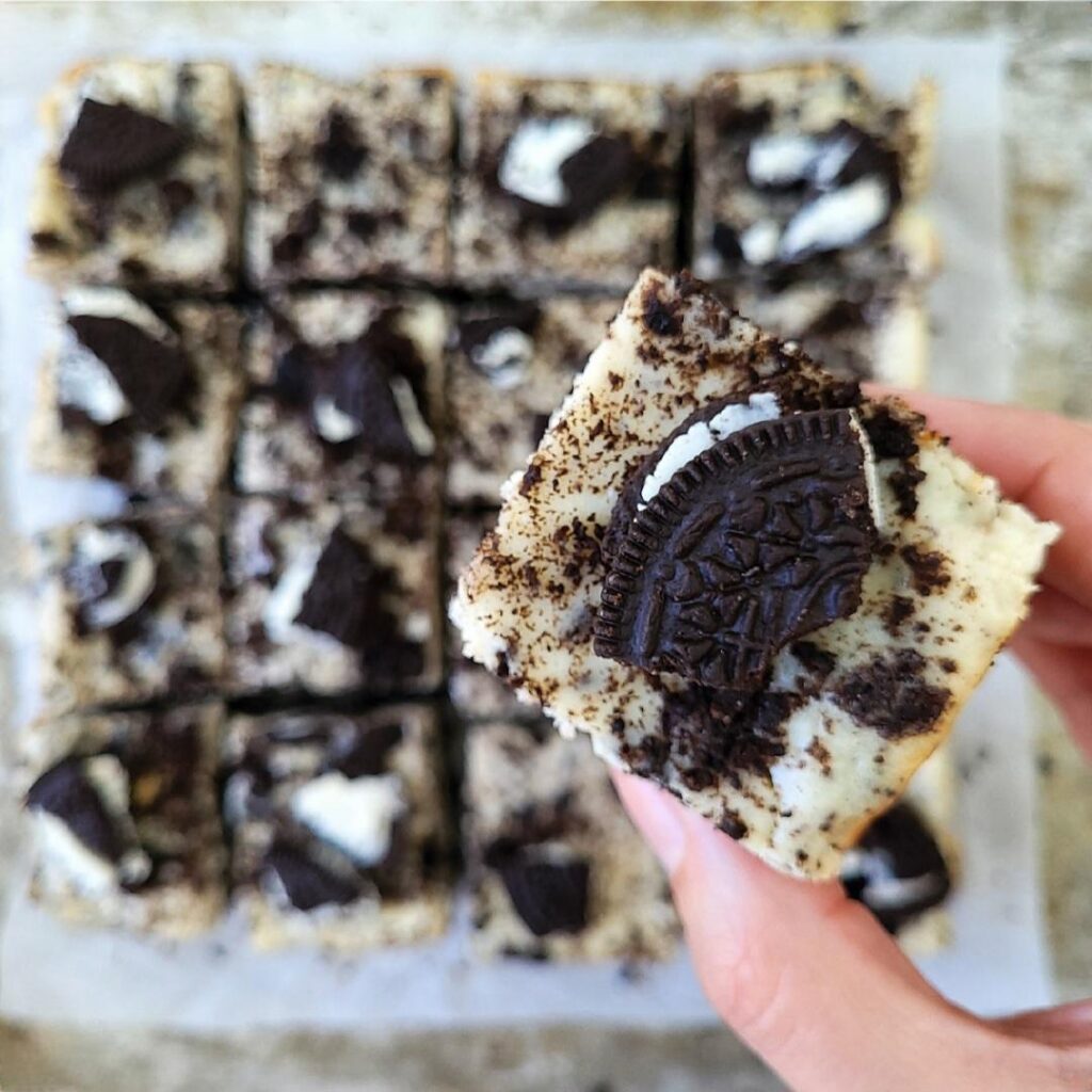 oreo cheesecake bars topped with broken Oreo cookie pieces. close up view of a hand holding a square cheesecake bar. 