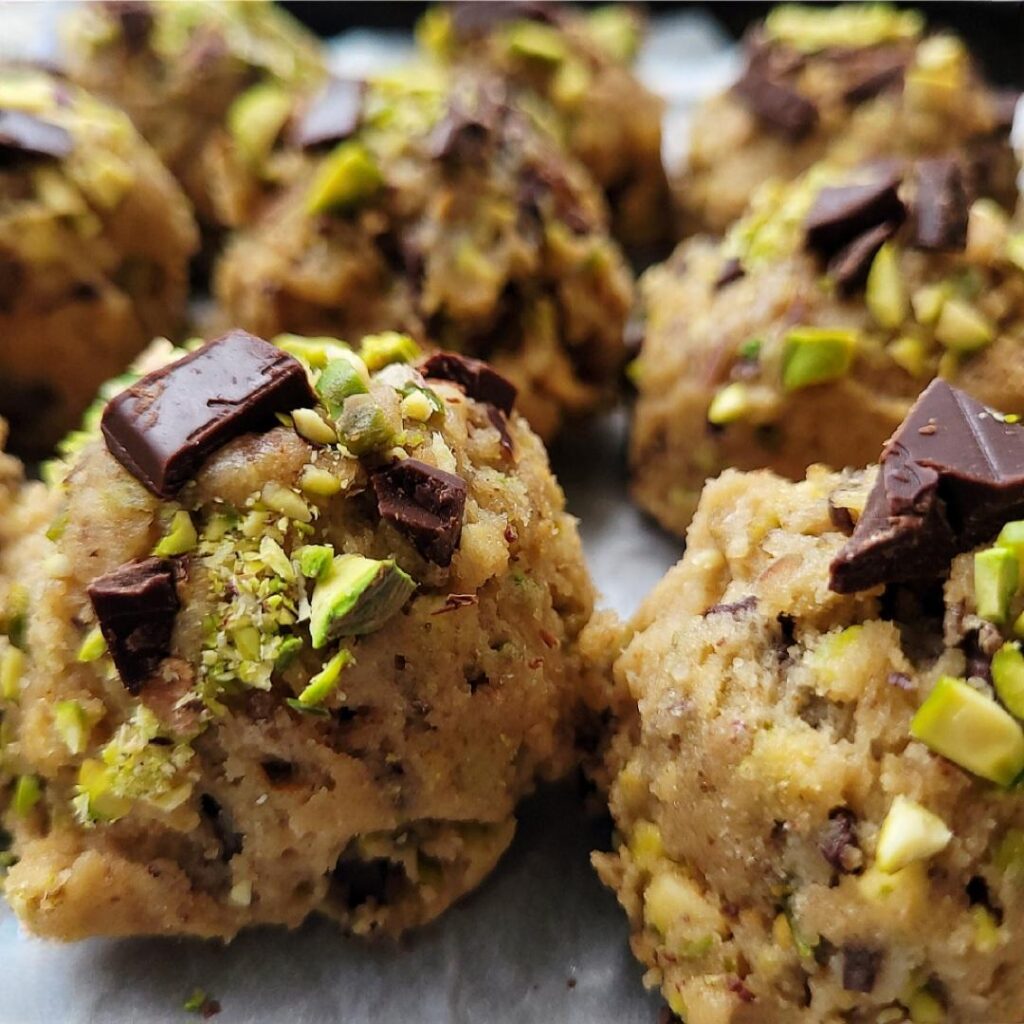 pistachio cookies. close up view of unbaked pistachio cookie dough balls topped with chopped pistachios and chunks of dark chocolate. 