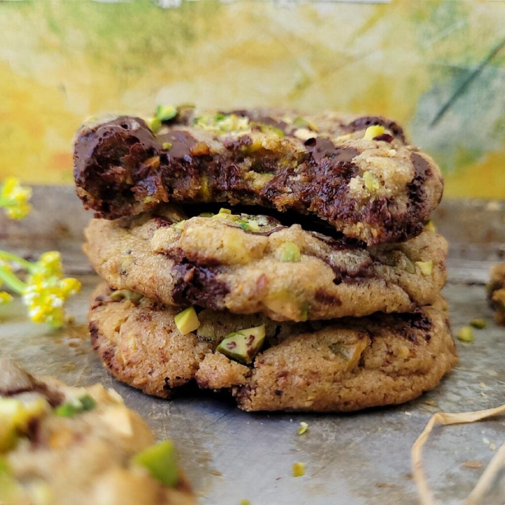 stack of three pistachio cookies with dark chocolate chunks. the top cookie has a bite missing so you can see the inner crumb, nuts and chocolate. 