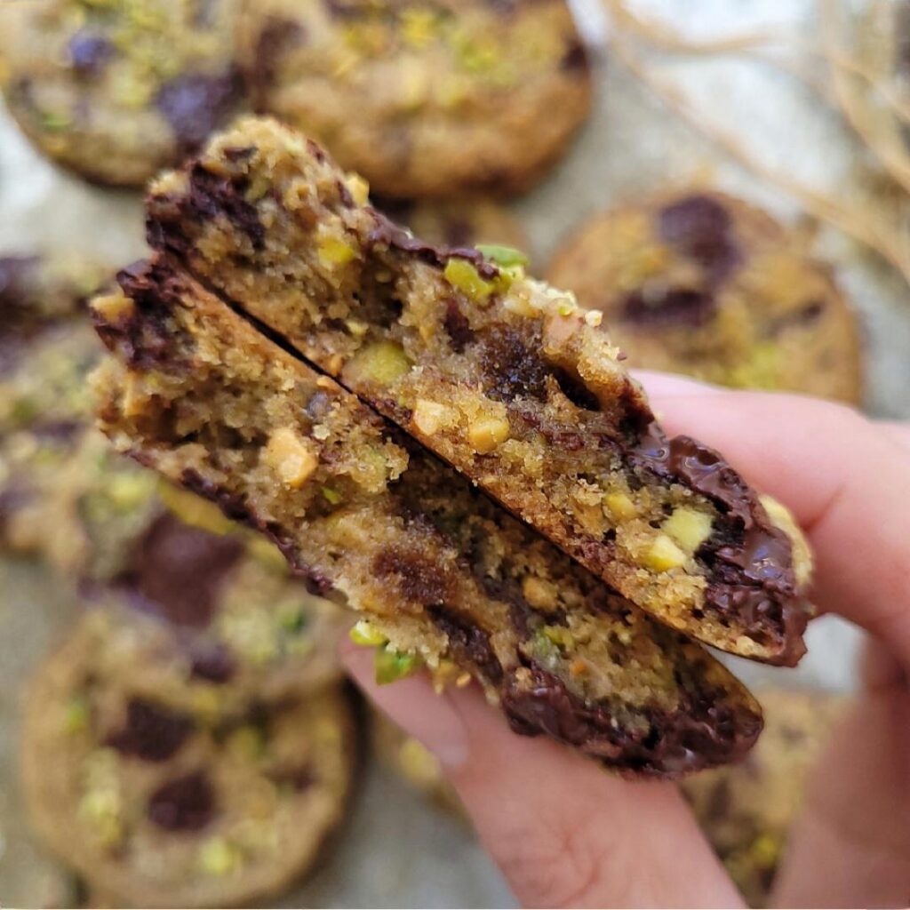 pistachio cookies with dark chocolate. close up view of a hand holding two halves of a broken cookie so you can see the inner crumb, chunks of chopped pistachios and melted dark chocolate. 