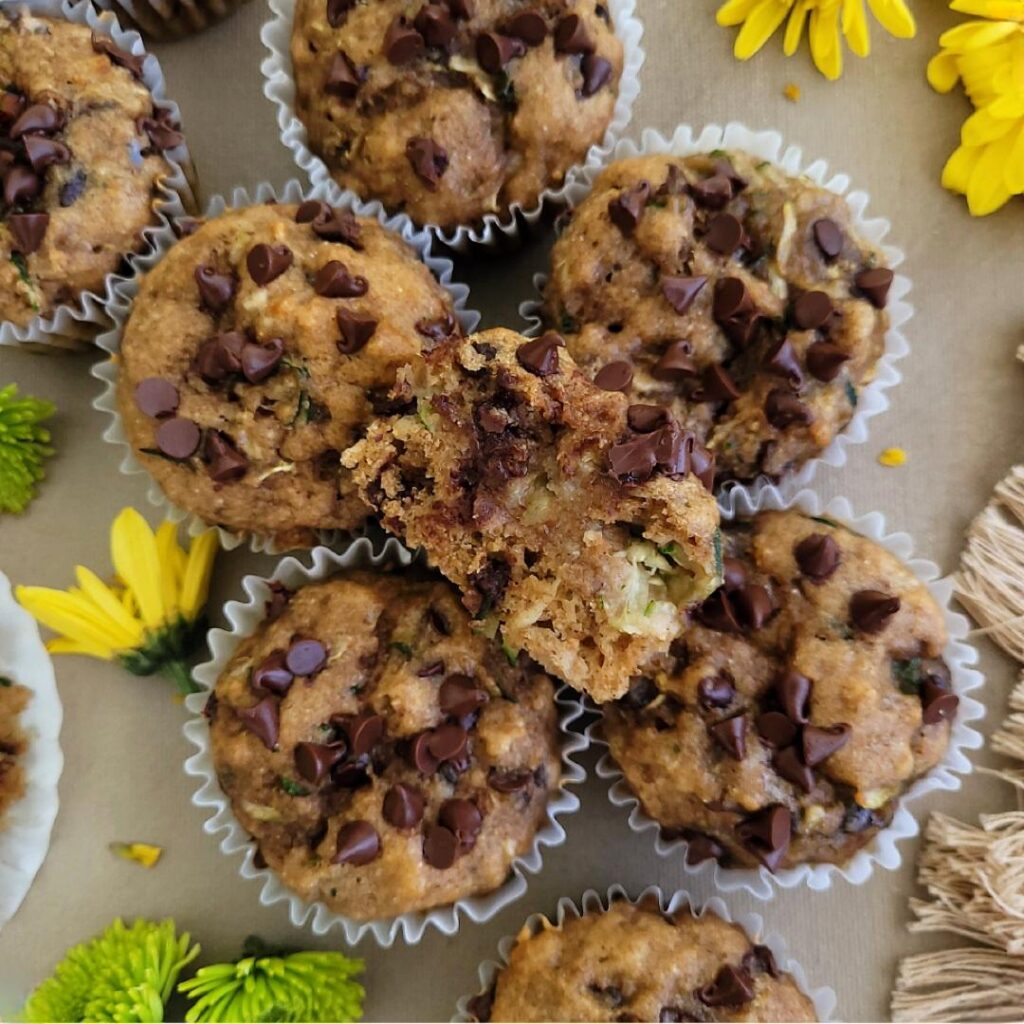 banana zucchini muffins. top down view of muffins in wrappers topped with mini chocolate chips. one muffin has been sliced in half and is facing upwards. 